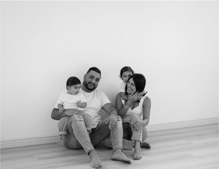 Black and white photo of a man with two children sitting together against a white wall, barefoot and smiling at the camera